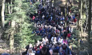 Tłum pielgrzymów podąża leśną ścieżką. Widać drewnianą wiatę i drzewa. Święty Krzyż trasa jest pełna ludzi.