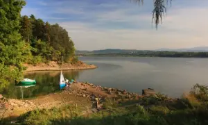 Spokojne jezioro żywieckie z łódkami przy brzegu, otoczone zielonymi drzewami i wzgórzami w oddali.