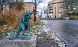 Mały smok z brązu, symbol smoczy szlak kraków, stoi na kamiennym postumencie. W tle miejski krajobraz z budynkami i drzewami.