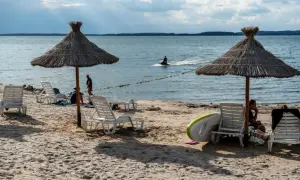 Słoneczne jezioro z plażą, leżakami i parasolami. Na wodzie widać skuter wodny.