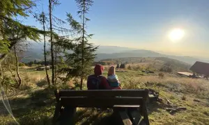 Para siedzi na ławce, podziwiając widok na góry. Słońce zachodzi nad horyzontem, malując niebo na złote barwy. To piękny widok z mogielicy z Jurkowa.