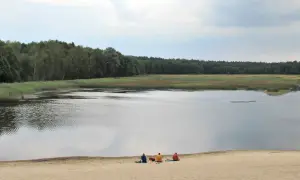 Trzy osoby i pies odpoczywają nad brzegiem czarnego stawu, otoczonego lasem.