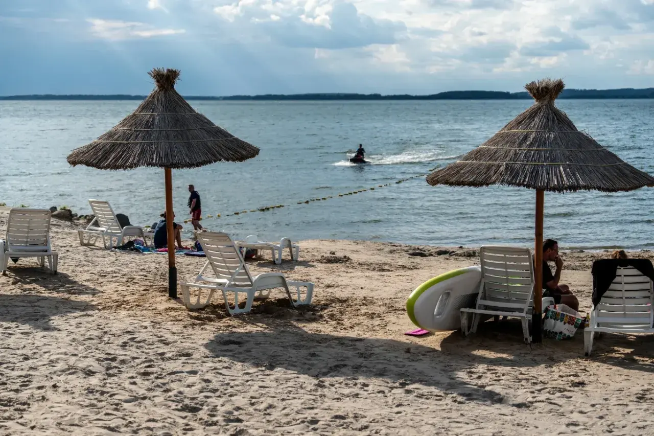Słoneczne jezioro z plażą, leżakami i parasolami. Na wodzie widać skuter wodny.