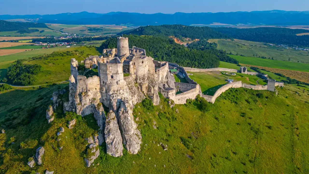 Majestatyczny zamek na skale, otoczony zielonymi wzgórzami i polami. Słoneczna Słowacja w pełnej krasie, zapraszająca do odkrywania historii.