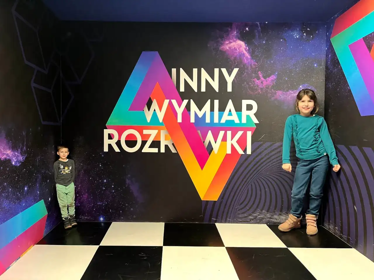 Dzieci w "Innym Wymiarze Rozrywki" w Warszawie. Kolorowa ściana z napisem i kosmicznym motywem tworzy niesamowite atrakcje dla dzieci.