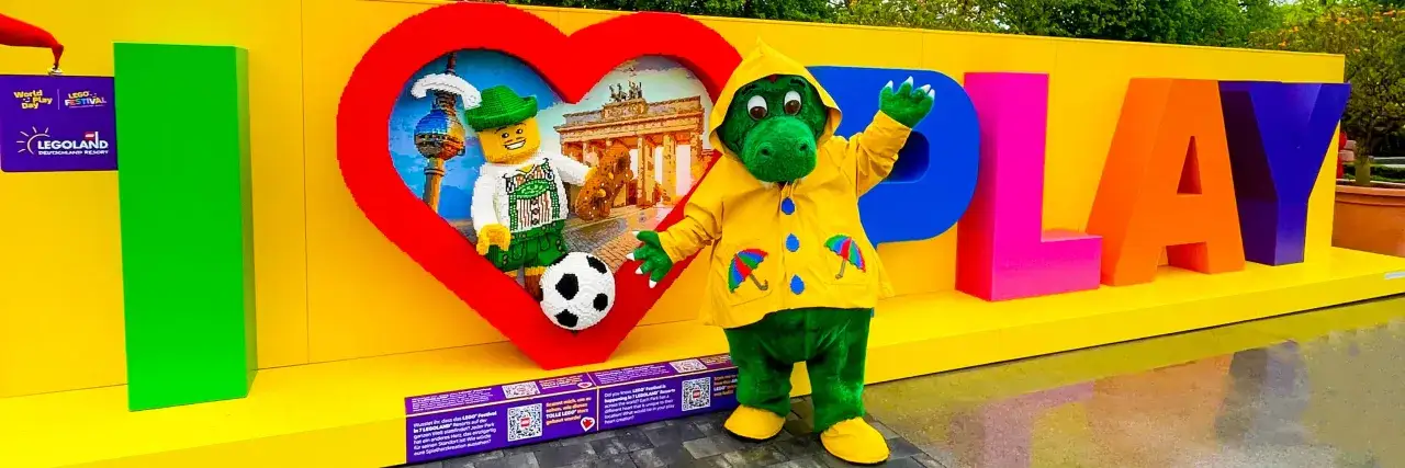 Maskotka w żółtej pelerynie i ludzik z klocków LEGO w sercu, na tle napisu 