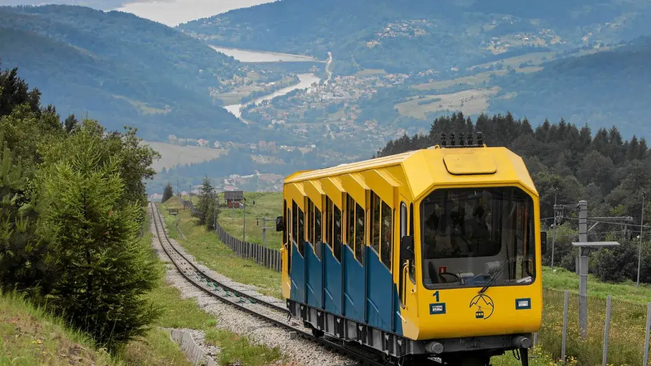 Żółty wagonik kolejki linowo-terenowej wjeżdża na górę Żar. Trasa kolejki prowadzi przez malownicze, zielone wzgórza.