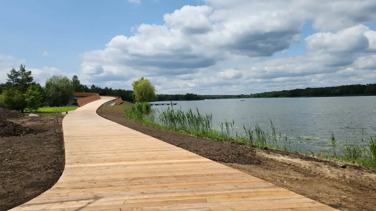 Drewniana kładka wije się nad brzegiem jeziora Paprocany. W oddali widać las i spokojną taflę wody.