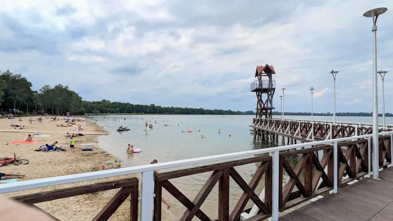 Plaża nad jeziorem, ludzie odpoczywają, pływają. Widoczna drewniana wieża ratownicza i pomost. Trasa Pogoria 3 tętni życiem.