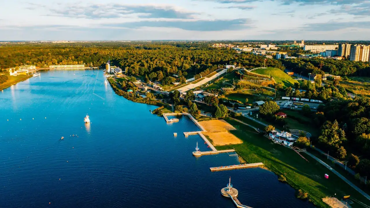 Słoneczne jezioro z piaszczystą plażą, pomostami i zielonymi terenami rekreacyjnymi, otoczone lasem i zabudowaniami miejskimi.