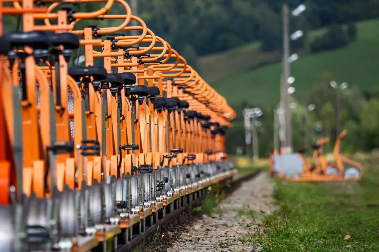 Pomarańczowe drezyny Bieszczady czekają na turystów. Rząd pojazdów na torach kolejowych, w tle zielone wzgórza.